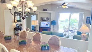 A dining area with a wooden table, chairs, and a chandelier. There is a television in the background, and a window with a view of the outdoors. - EP1051SEL - Private Vacation Pool Home Near Disney (Kissimmee)