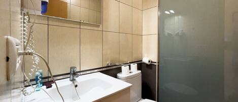 Un baño con lavamanos blanco, espejo y inodoro. Hay un secador de pelo en la pared y una cortina de ducha.