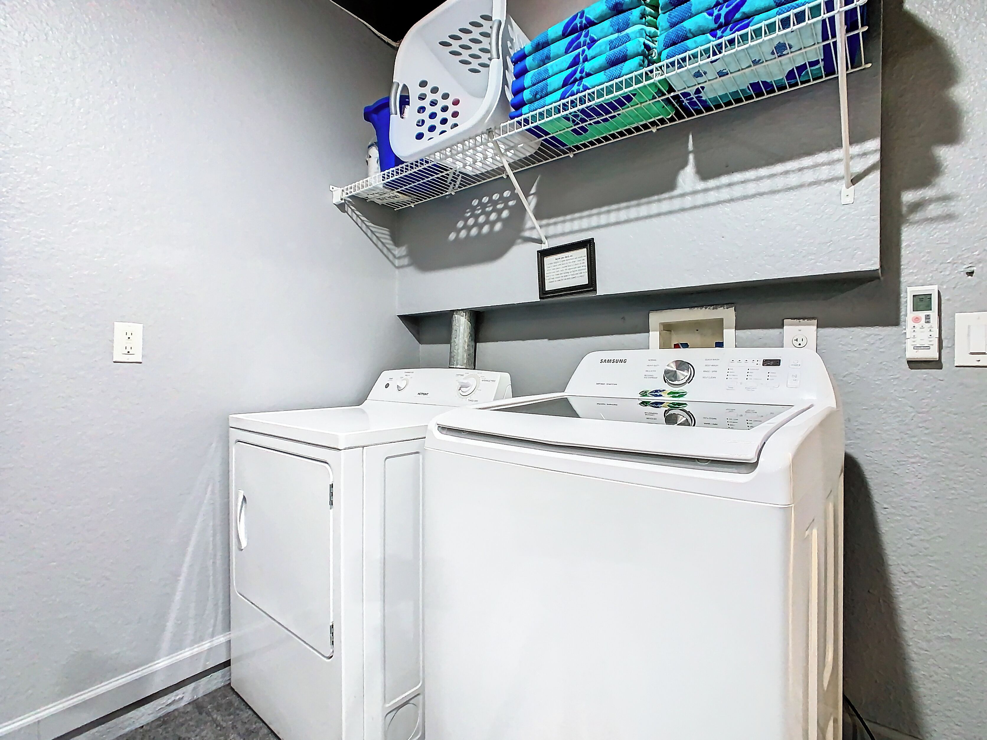 A laundry area with two white front-loading washers and dryers, a metal shelf above holding folded towels, and a wall-mounted thermostat.