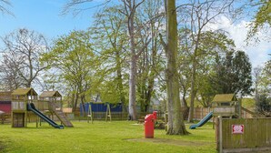 Une aire de jeux avec des toboggans et des structures d’escalade, entourée d’arbres et délimitée par une clôture.