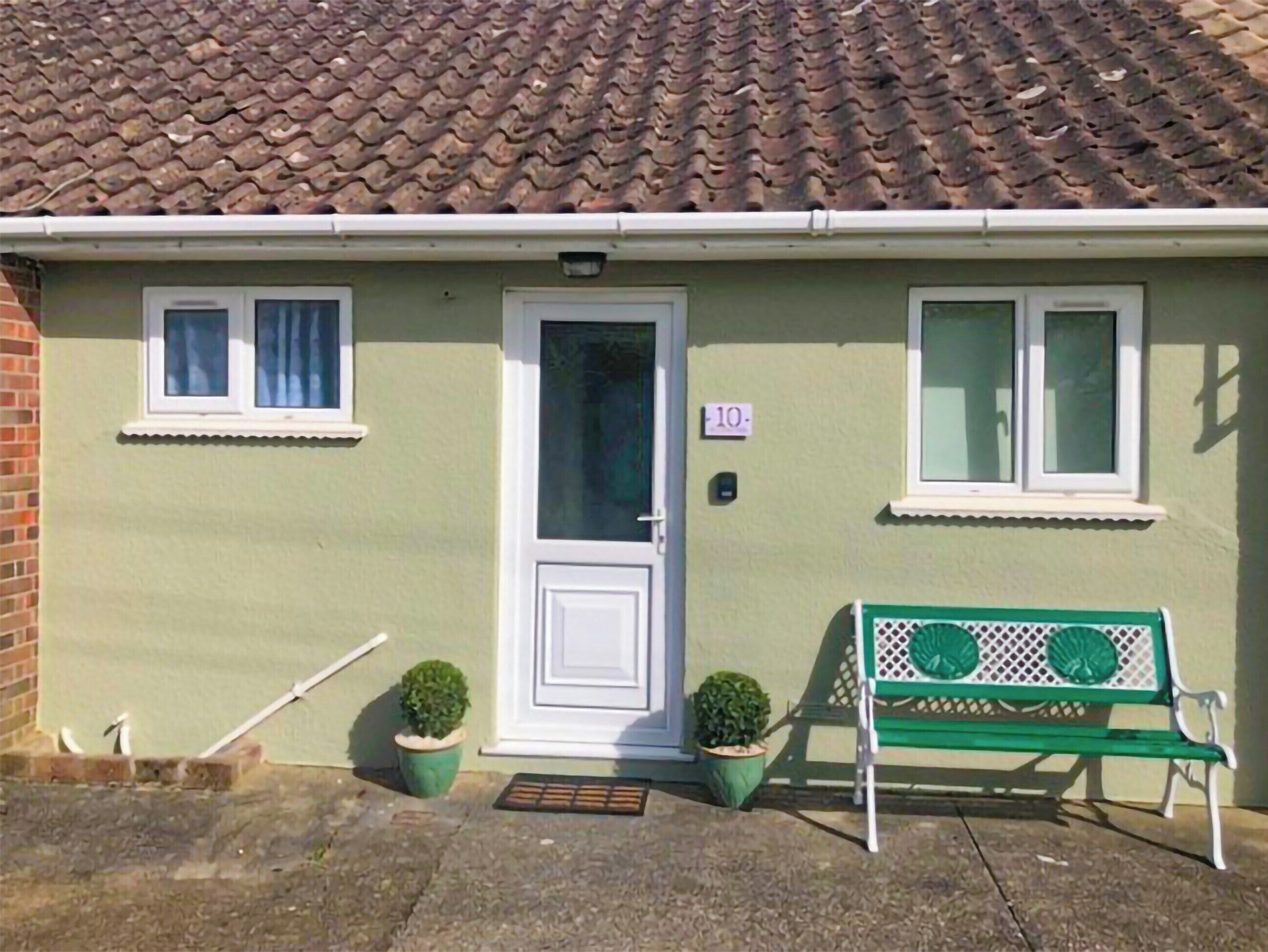 A single-story house with a tiled roof, a front door, two windows, and a green bench outside.