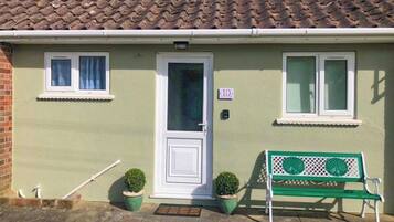 A single-story house with a tiled roof, a front door, two windows, and a green bench outside.