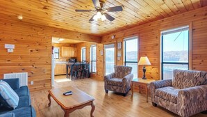 A spacious living room with wooden walls and ceiling, a ceiling fan, a sofa, a coffee table, and a view of the kitchen and outside.