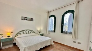 A bedroom with a bed, a nightstand, a lamp, and two windows with curtains. - Residence Skipper (San Teodoro)