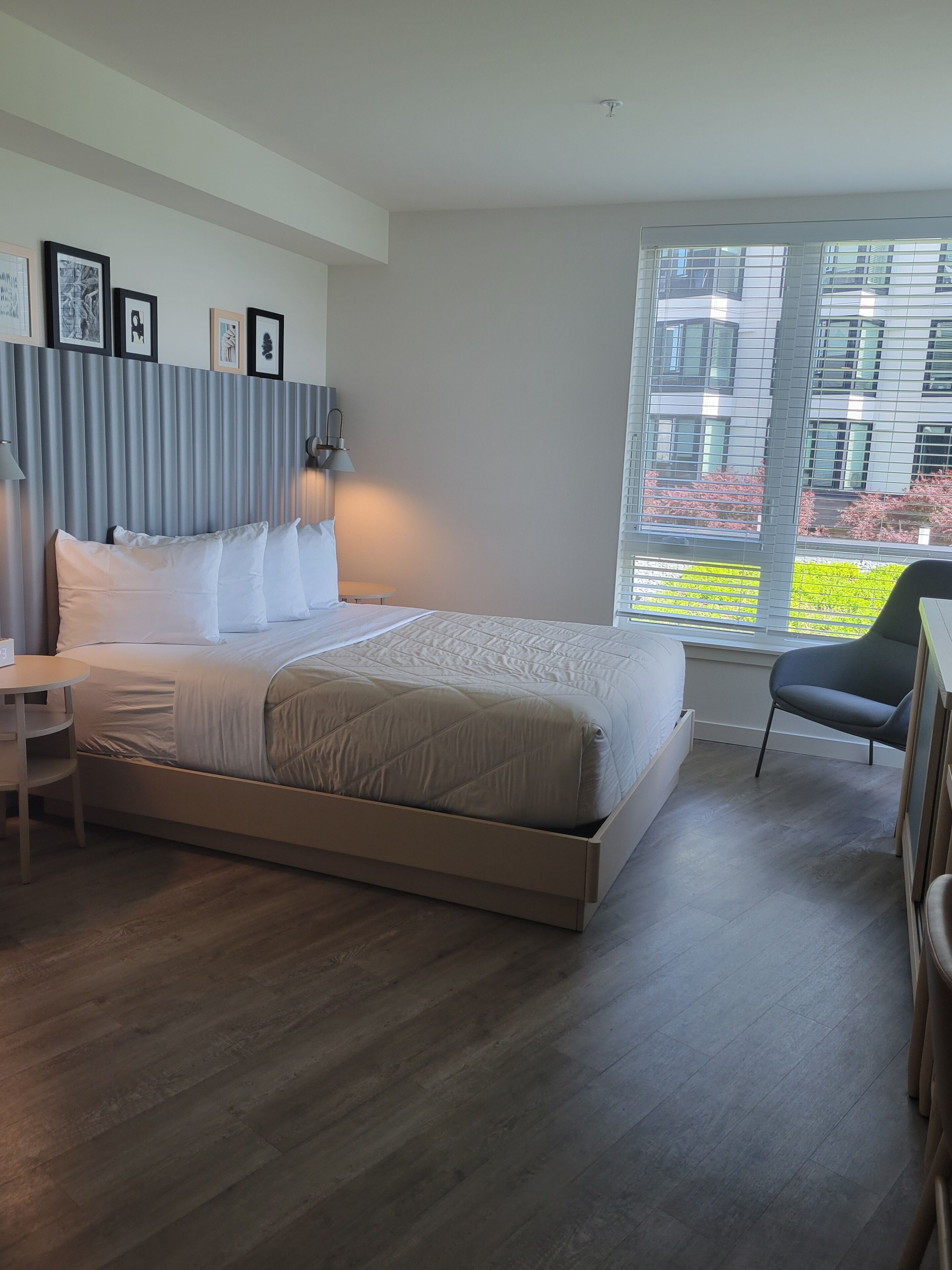 Quarto de hotel moderno com uma cama grande, mesa de cabeceira, cadeira, janela com vista para edifícios e quadros nas paredes.