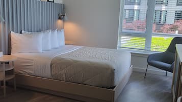 A modern hotel room with a large bed, a bedside table, a chair, a window with a view of buildings, and framed pictures on the wall.