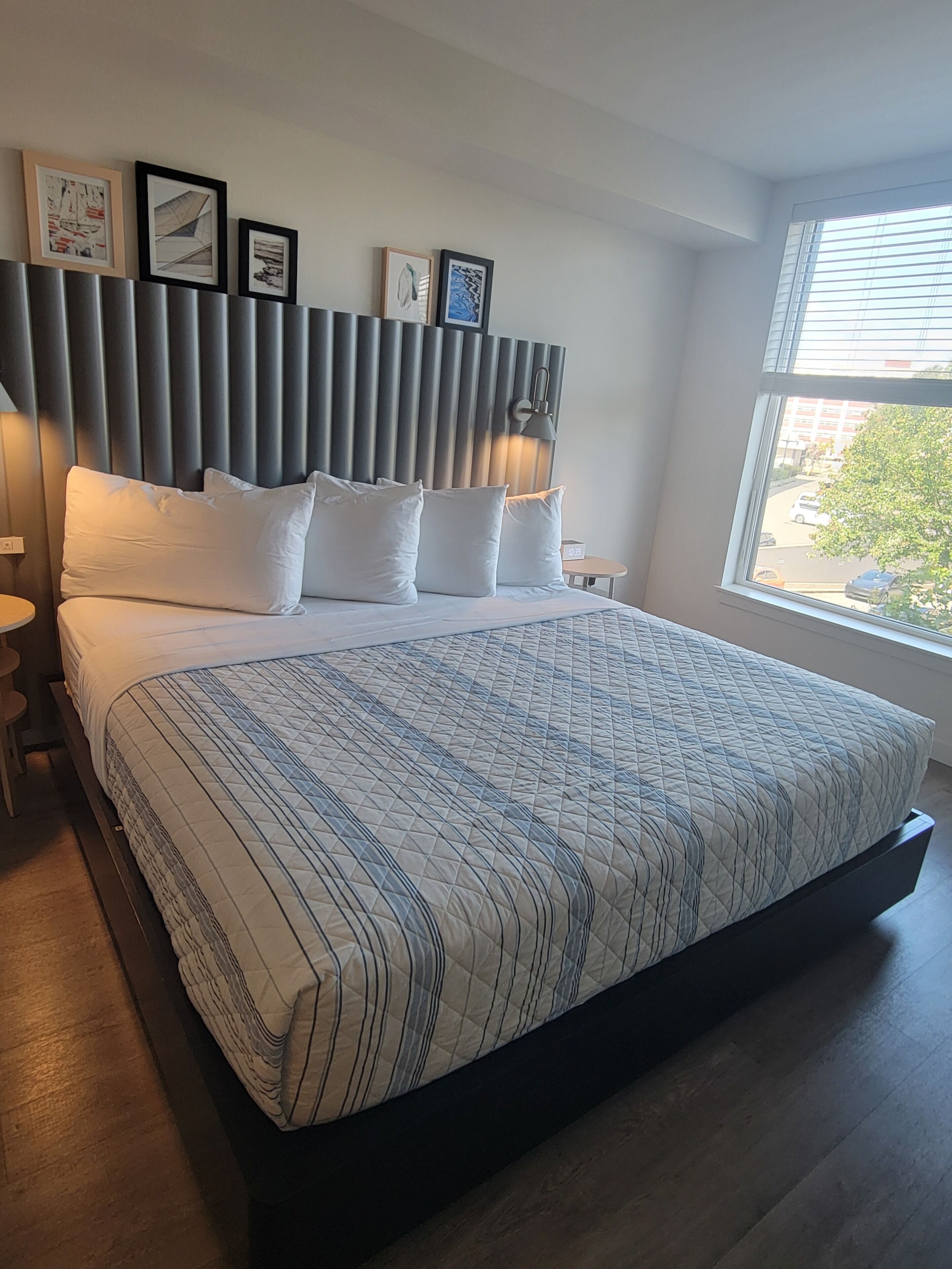 A modern hotel room with a large bed, a headboard with vertical slats, a bedside table, a nightstand, and a window with a view of the outside.
