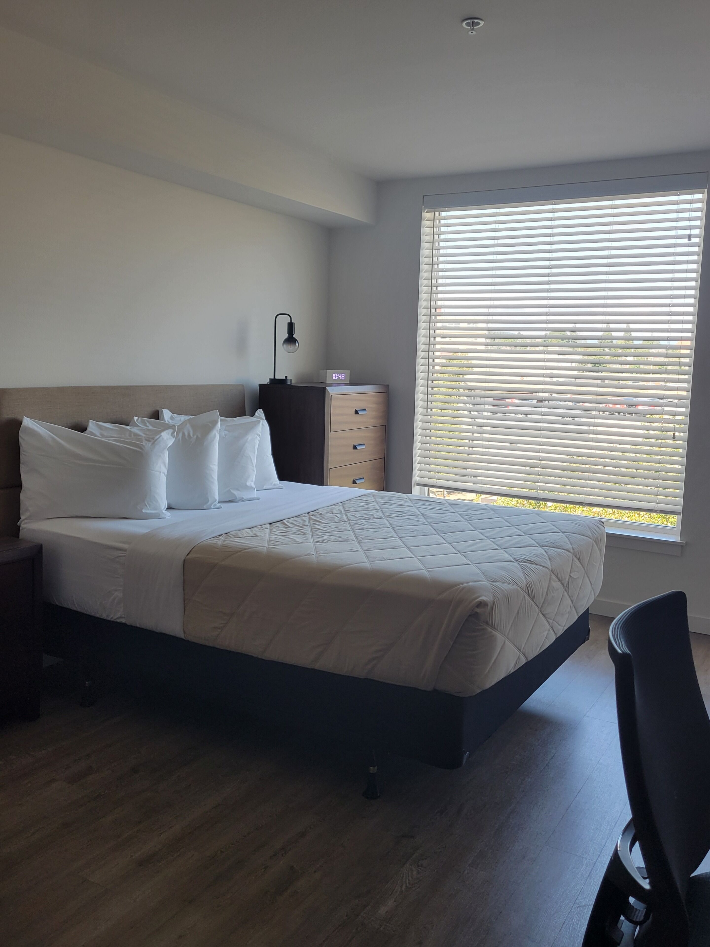Quarto com cama, mesa de cabeceira, luminária, relógio e janela com persianas.