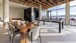 A spacious indoor area with a pool table, dining table, and chairs, large windows offering a view of the city. - Marina Square Suites (Bremerton)