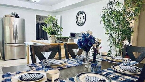 Un comedor con una mesa de madera dispuesta para cuatro personas, rodeada de sillas. Hay dos plantas en macetas, un reloj en la pared y un refrigerador de acero inoxidable al fondo.