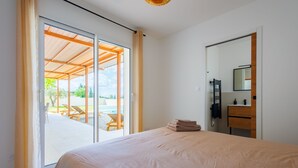 Een slaapkamer met een groot bed, een schuifdeur naar een buitengebied met een zwembad en ligbedden, een houten plafond en een kleine badkamer met spiegel en wastafel.