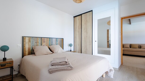 Een slaapkamer met een groot bed, een houten hoofdbord, een nachtkastje met een lamp en uitzicht op een zithoek.