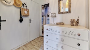 Um corredor com um aparador branco com a etiqueta "Deauville", um espelho e dois chapéus pendurados na parede.