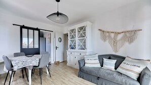 Uma sala de estar moderna com um sofá cinzento, uma mesa de jantar com cadeiras cinzas, um armário branco e um painel de macramé na parede.