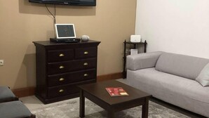 A living room with a flat-screen TV mounted on the wall, a dark wooden cabinet with drawers, a grey sofa, a small table with a magazine, and a patterned rug.