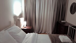 A hotel room with a bed, a bedside table with a lamp, a dresser, and a window with curtains.