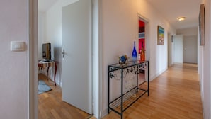 Um corredor com piso de madeira, uma mesa de apoio de vidro com objetos decorativos e uma porta que dá para outro quarto.