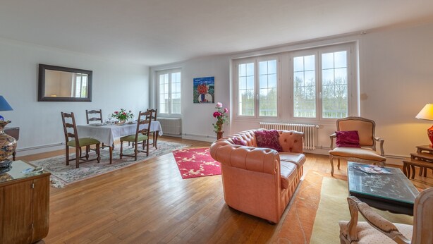 Uma sala de estar espaçosa com piso de madeira, um sofá rosa, área de jantar com mesa e cadeiras, e grandes janelas que oferecem iluminação natural.