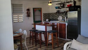A compact kitchen with a refrigerator, microwave, and tiled backsplash. There is a dining area with a table and chairs, and a bar stool area.