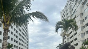 A gated entrance with a stone lion statue, palm trees, and a modern building in the background. - Acogedora casa dentro de fraccionamiento a orilla de la playa (Mazatlán)