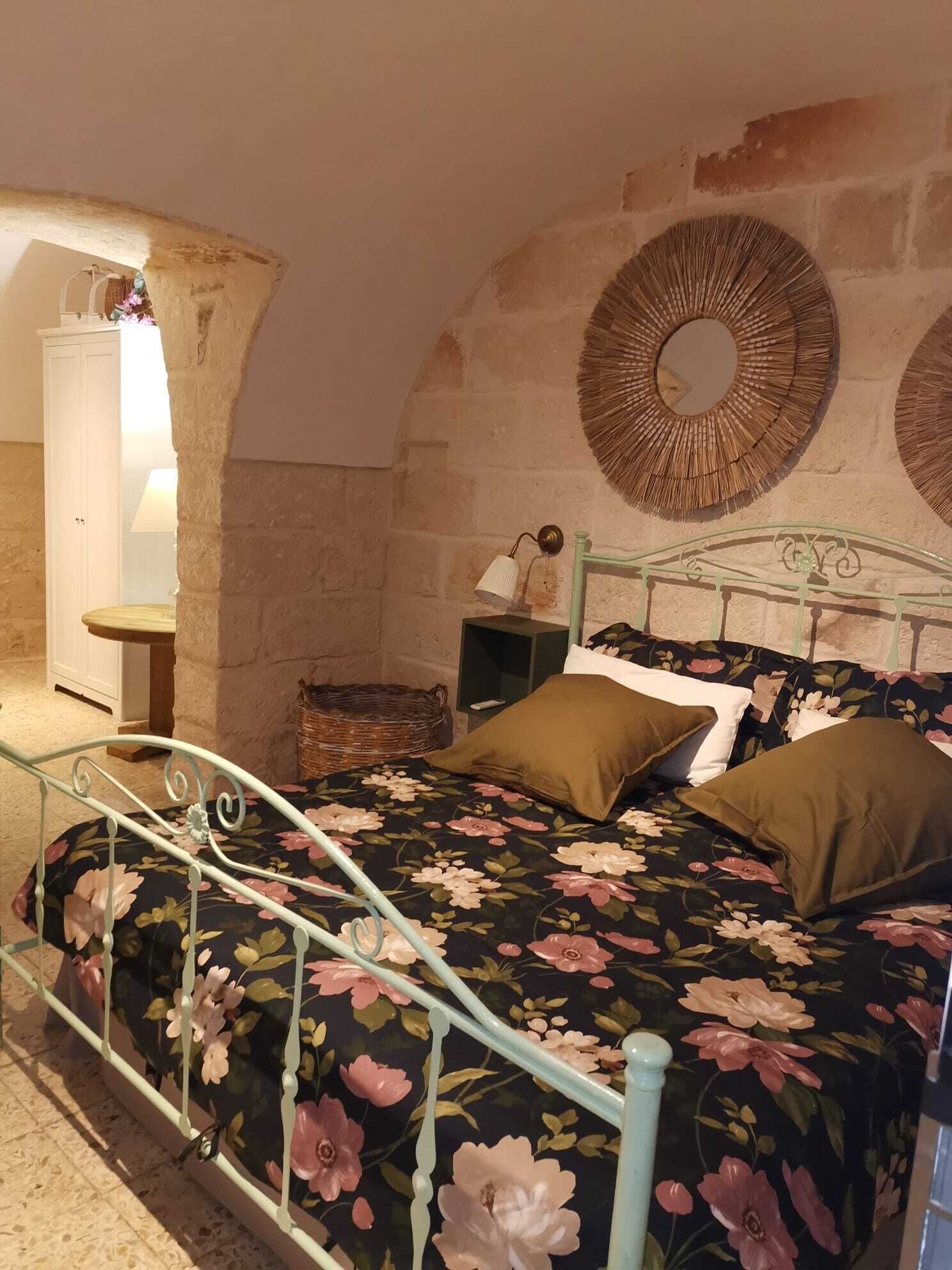 Ein Schlafzimmer mit floralem Bettbezug, einem Metallbett, einem Nachttisch mit Lampe und einer Steinwand mit rundem Spiegel.