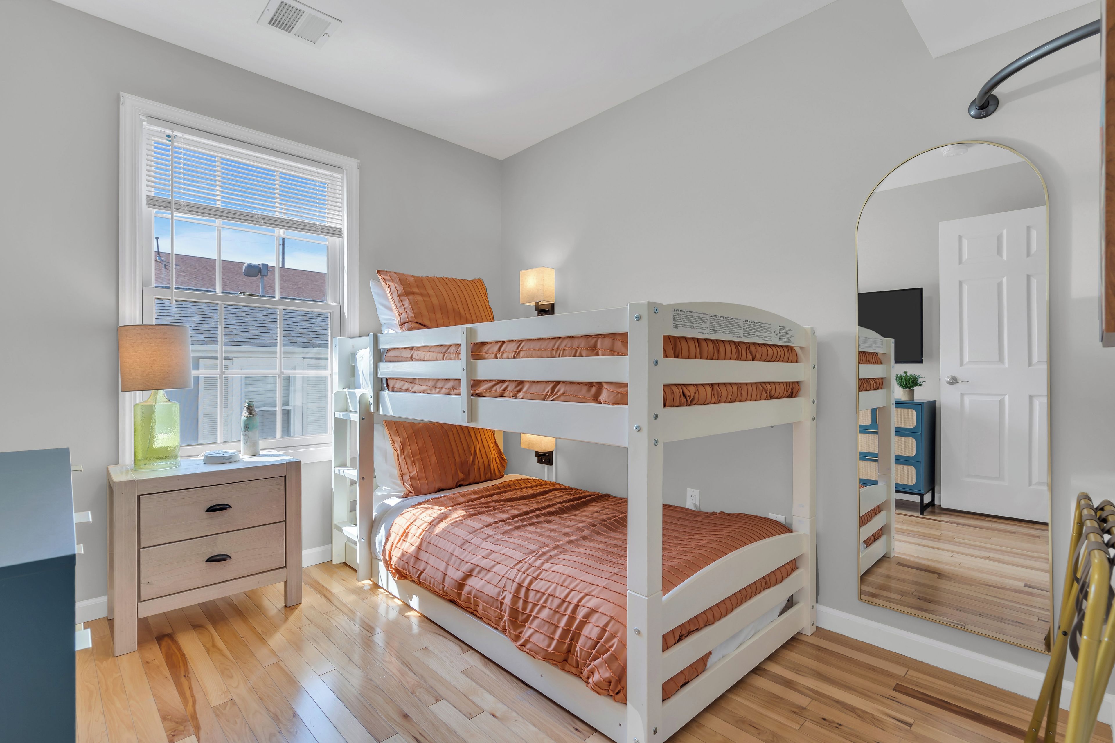 A bunk bed with a bedside table and lamp, a window with blinds, and a mirror on the wall.