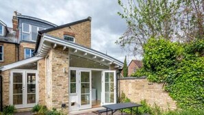Une maison à deux étages, avec une façade en briques, un porche à l’avant et un petit jardin agrémenté d’une table et de bancs.