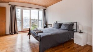 Une chambre avec un plafond en bois, un lit avec une literie grise, une table de chevet avec une lampe, et une vue dégagée sur l’extérieur grâce à une grande fenêtre.
