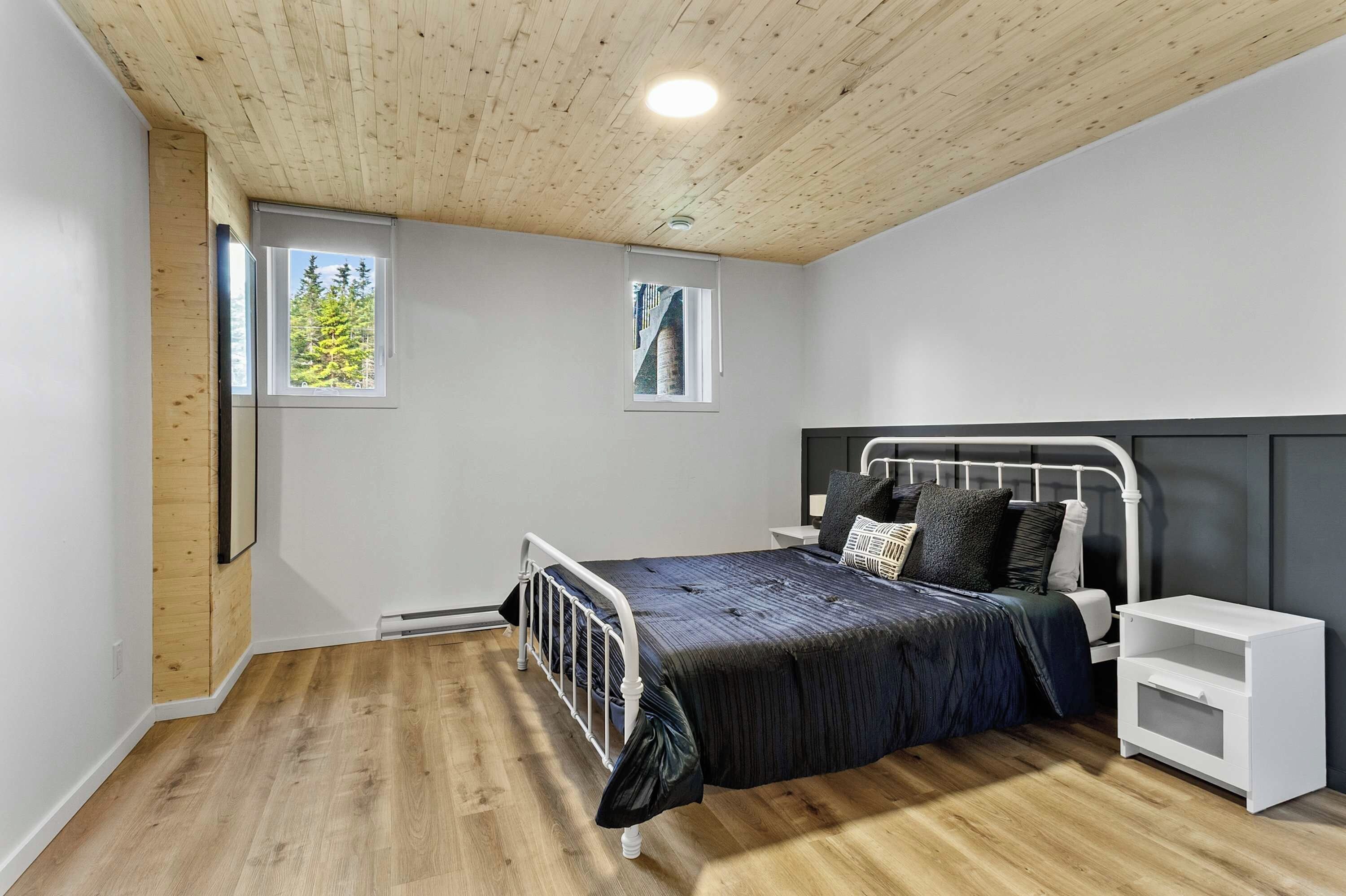 A bedroom with a bed, bedside tables, and two windows with views of trees.