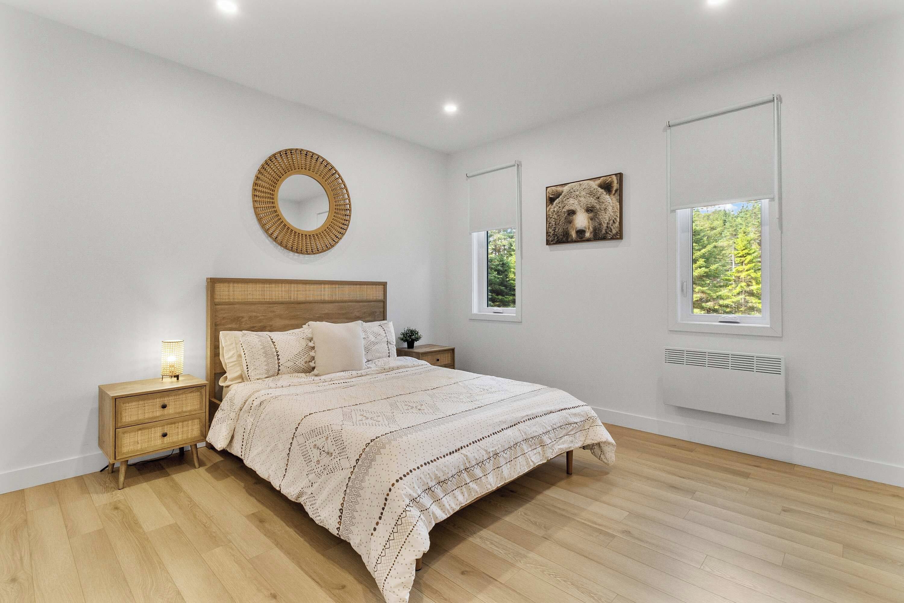 A bedroom with a wooden bed, a bedside table with a lamp, a round mirror, and two windows with views of greenery.