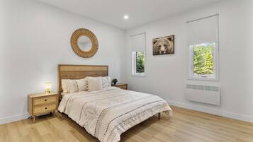 A bedroom with a wooden bed, a bedside table with a lamp, a round mirror, and two windows with views of greenery.