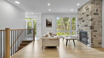 Un salon moderne avec une cheminée en pierre, un canapé, une table basse et de grandes fenêtres donnant sur une forêt.
