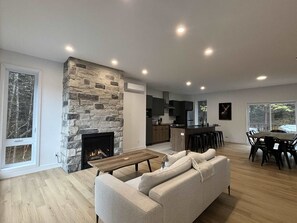 A modern living room with a stone fireplace, a sofa, a dining area with a table and chairs, and a kitchen in the background.
