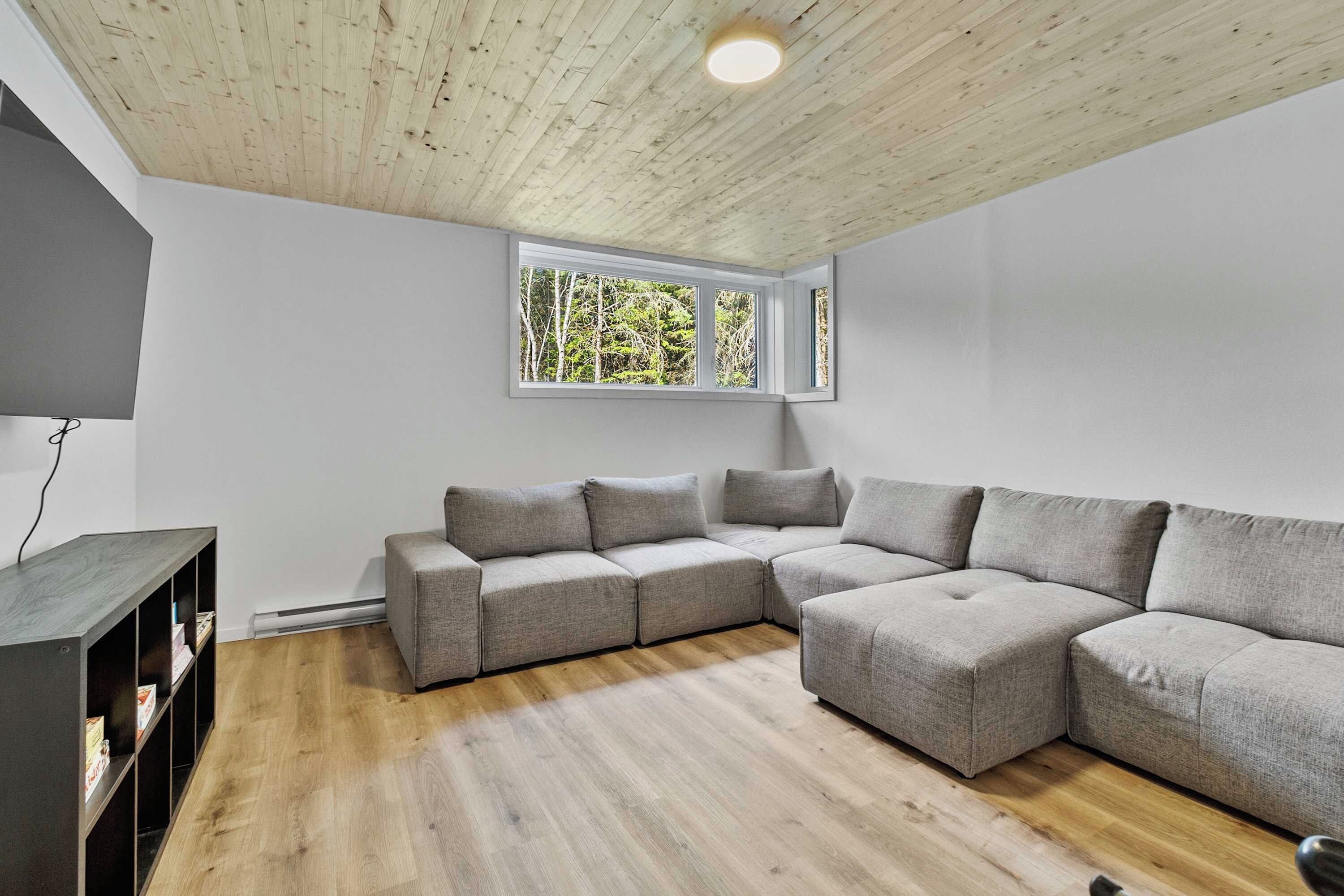 Un salon moderne avec un canapé d’angle, un plafond en bois et une fenêtre donnant sur la forêt.