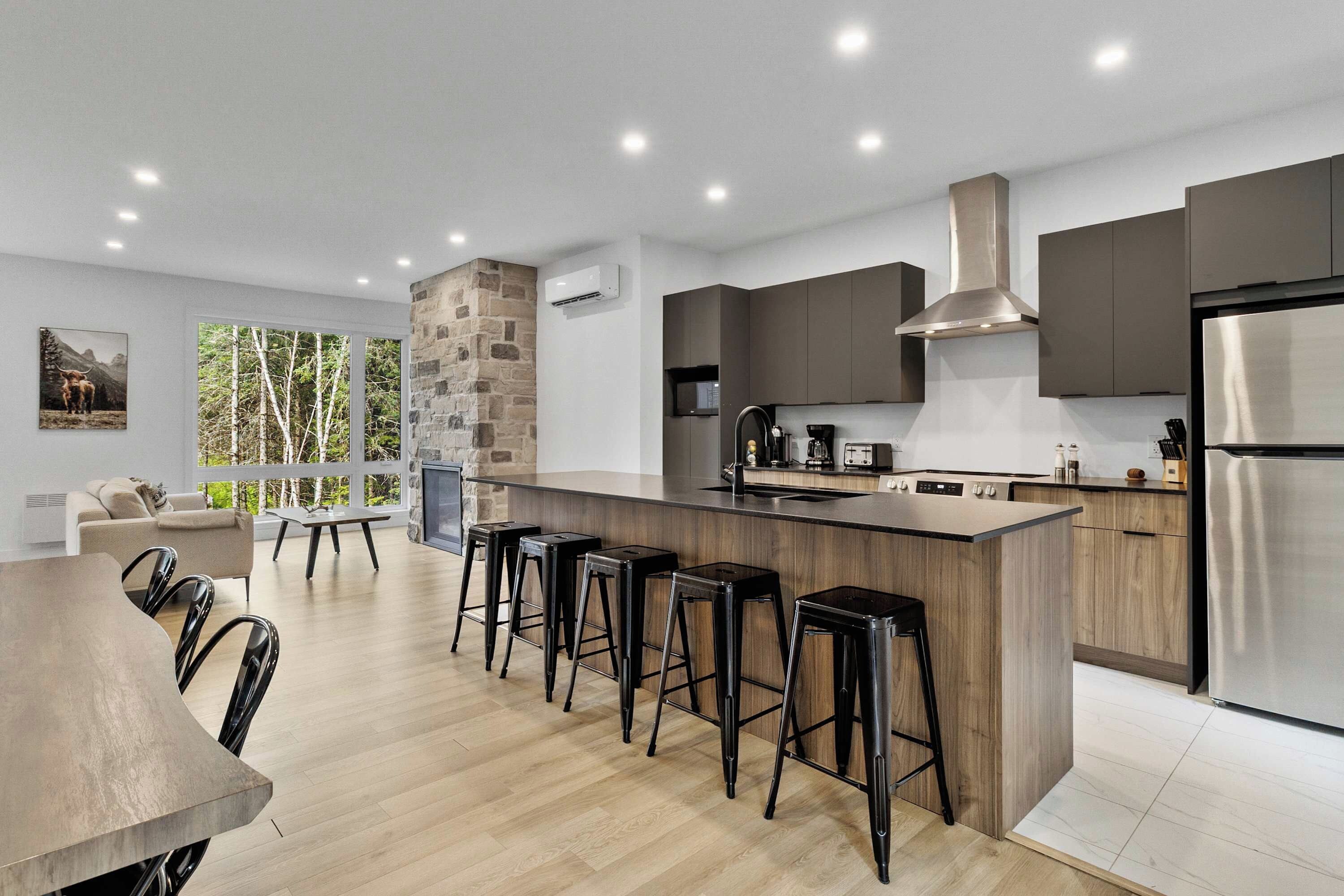 A modern kitchen with a central island, bar stools, and a dining area with a view of trees.