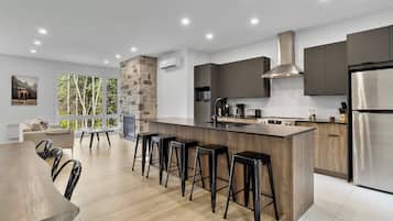 A modern kitchen with a central island, bar stools, and a dining area with a view of trees.