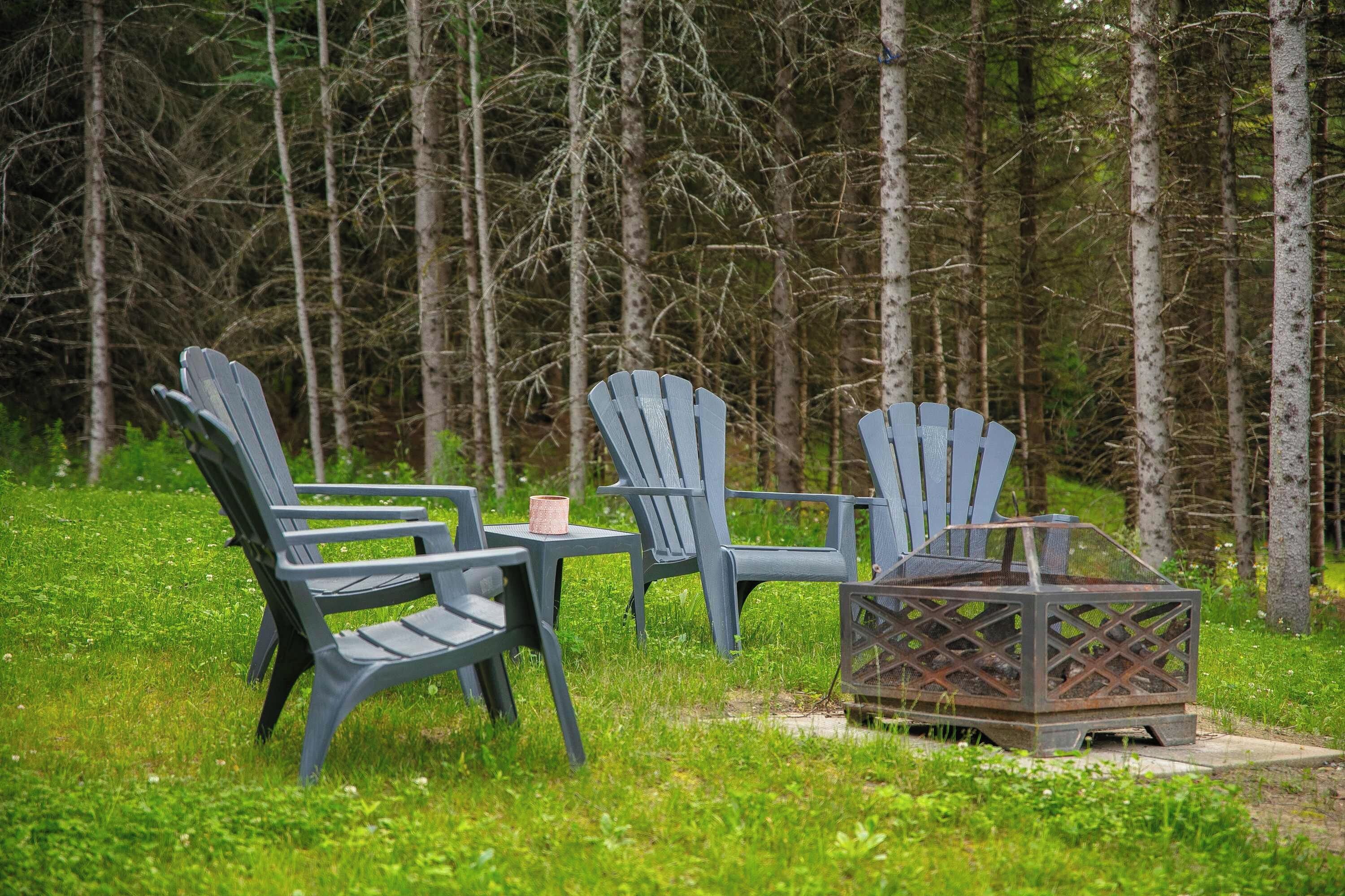 Two Adirondack chairs, a metal fire pit, and a small table with a cup, set in a grassy area with trees in the background.