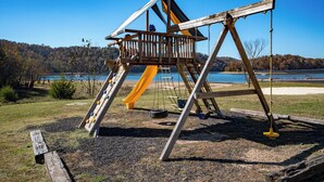 A wooden swing set with a slide, surrounded by a gravel area and a view of a lake and forest. - Chalet with 2 Bedrooms lakeside to Dale Hollow Lake #17 (Byrdstown)