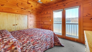Une pièce de cabane en bois comprenant un lit, un ventilateur de plafond et une grande fenêtre donnant sur une étendue d’eau.