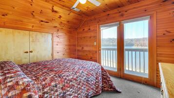 Puinen mökkimäinen huone, jossa on sänky, kattotuuletin ja suuri ikkuna, josta avautuu näkymä vesistölle.