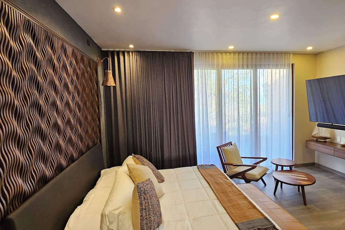 Une chambre d’hôtel moderne dotée d’un grand lit, d’un téléviseur fixé au mur, d’un coin salon avec un fauteuil et une petite table, ainsi que d’un panneau décoratif mural.