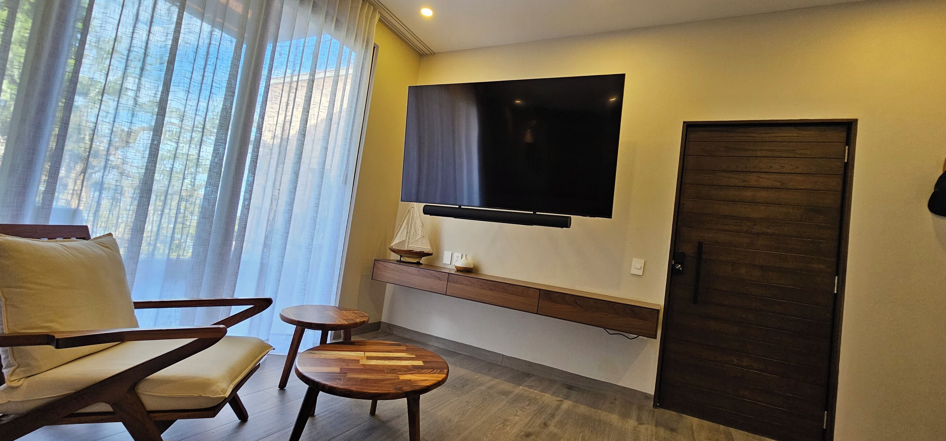 A modern room with a sofa, two small tables, a TV mounted on the wall, a wooden door, and a window with sheer curtains.