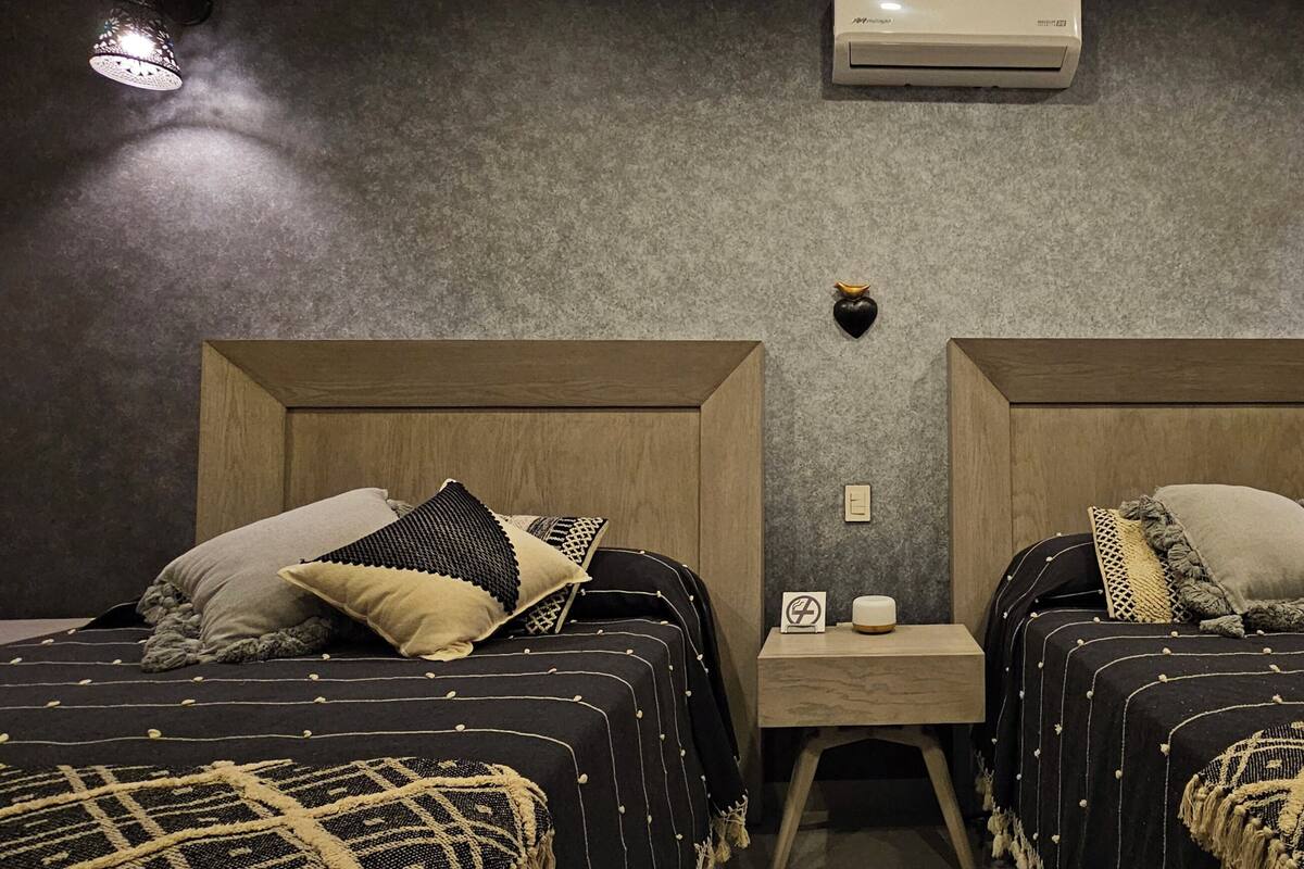 Une chambre d’hôtel avec deux lits, chacun agrémenté de draps et d’oreillers à motifs. Une petite table se trouve entre les lits, sur laquelle sont posés une bougie et un panneau.