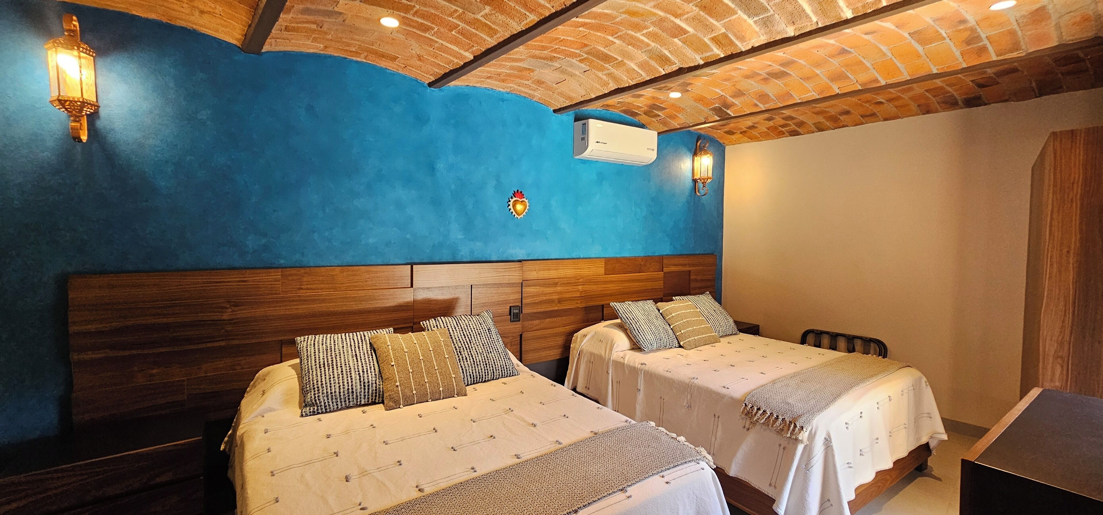 A room with two beds, a wooden headboard, a blue accent wall, and a brick ceiling.