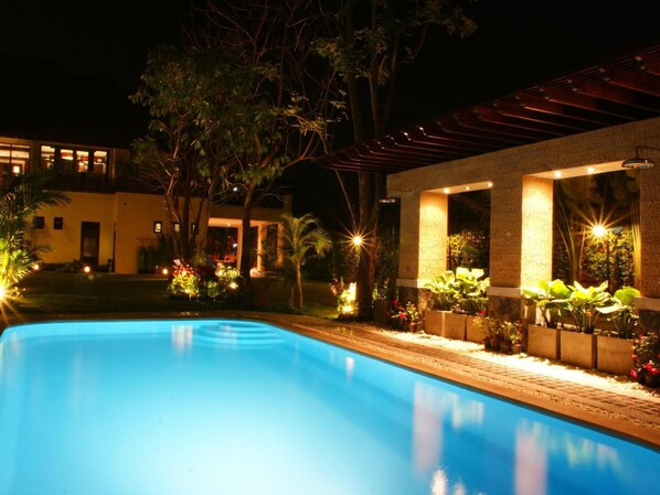 Une piscine nocturne, entourée de plantes, et un bâtiment dont les fenêtres sont illuminées.