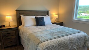 Uma cama bem arrumada com colcha estampada e almofadas, duas luminárias de cabeceira e uma mesa de cabeceira de madeira com gaveta.