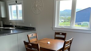 Uma área de refeições com mesa e cadeiras de madeira, uma cozinha com pia e bancada, um enfeite de parede com bússola e uma janela com vista para vegetação.