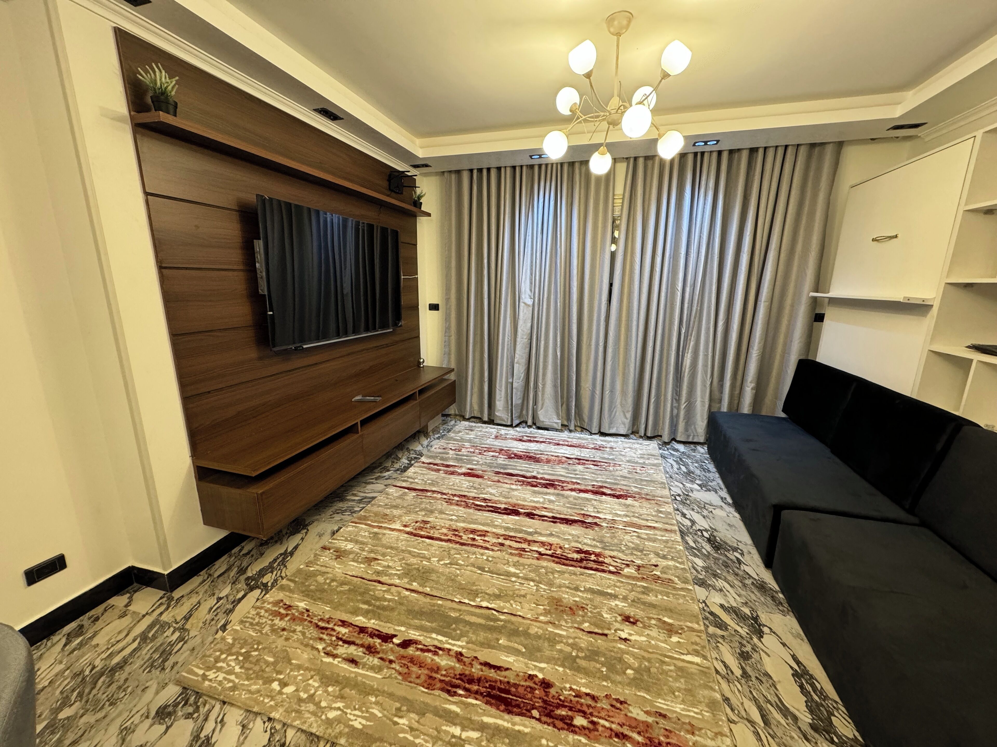 A modern living room with a flat-screen TV, a black sofa, a patterned rug, and a chandelier.