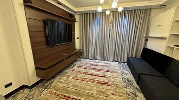 A modern living room with a flat-screen TV, a black sofa, a patterned rug, and a chandelier.