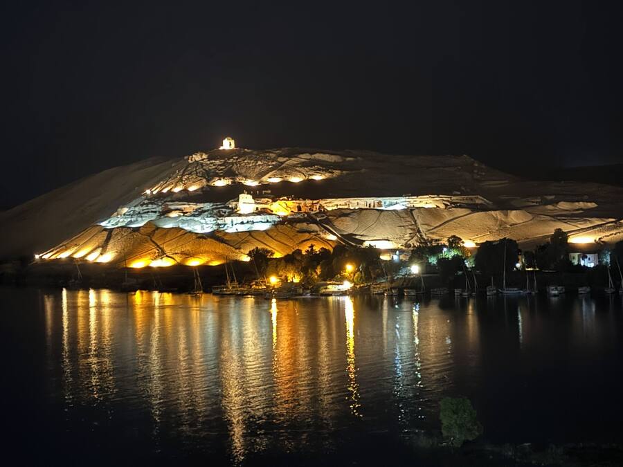 Un grand bâtiment illuminé, doté d’une structure centrale imposante et entouré de plusieurs bâtiments plus petits, situé au bord d’une étendue d’eau qui reflète les lumières.